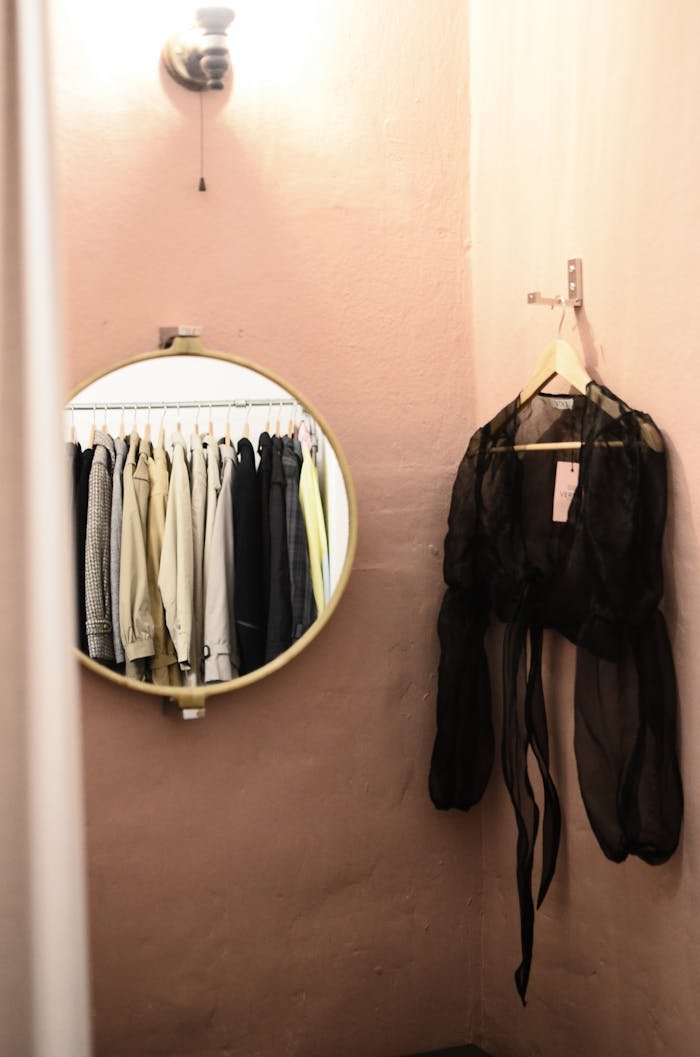 Interior of minimalistic room with lamp on wall and mirror with reflection of different clothes near blouse on hanger